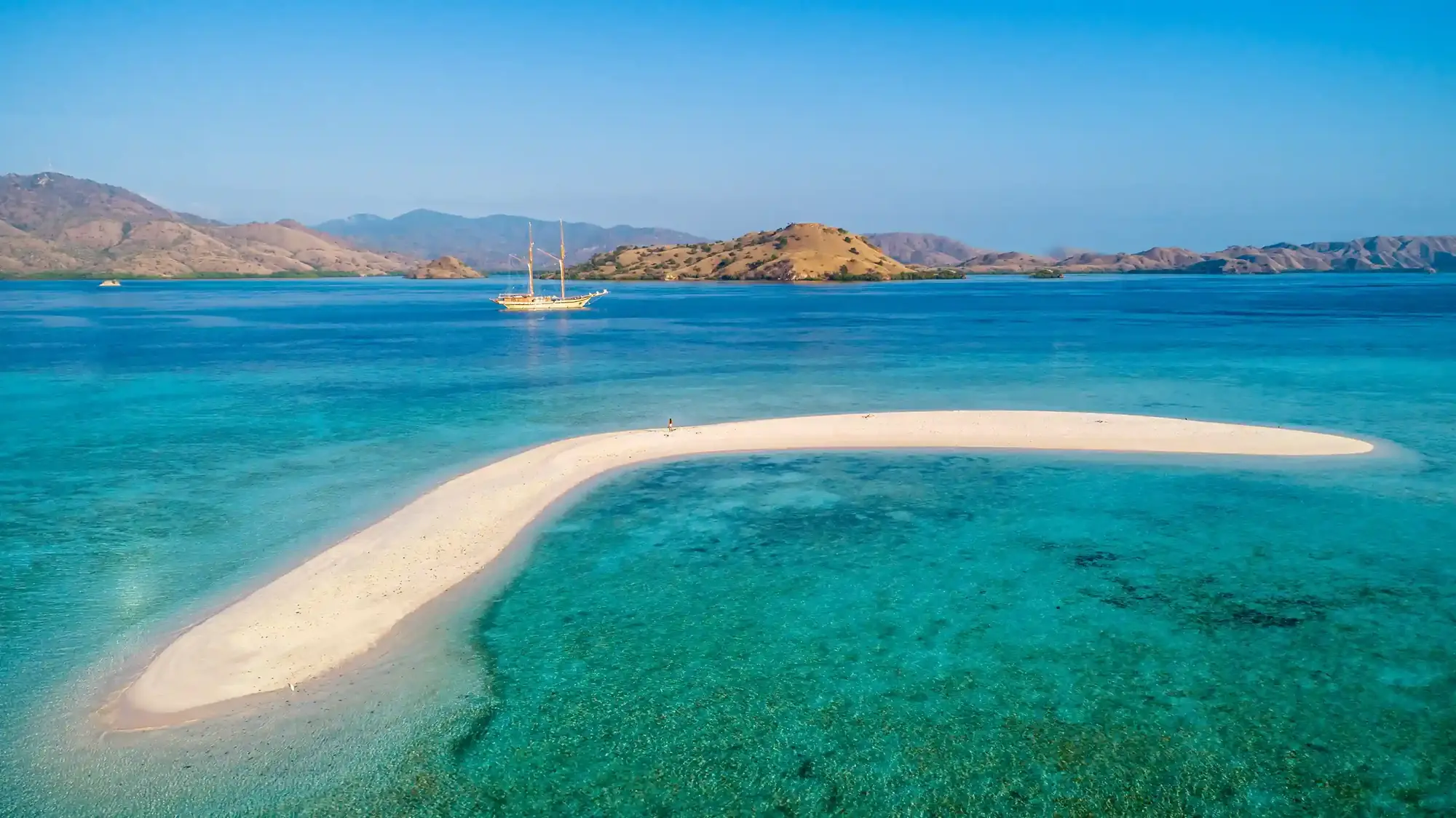 White sandbar curving through turquoise shallows with a sailing boat beyond